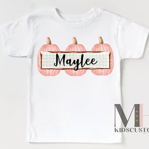 Puede incluir: Camiseta blanca con tres calabazas rosas y tallos marrones. El nombre "Maylee" est&aacute; escrito en escritura negra en una pancarta rectangular entre las calabazas. La camiseta tiene un logotipo de KidsCustom en la parte inferior.