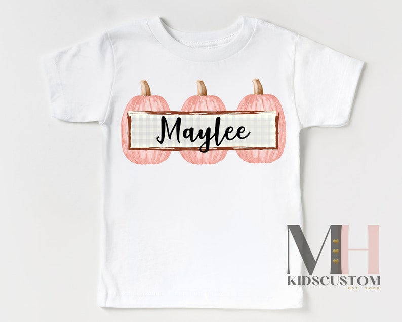 Puede incluir: Camiseta blanca con tres calabazas rosas y tallos marrones. El nombre "Maylee" est&aacute; escrito en escritura negra en una pancarta rectangular entre las calabazas. La camiseta tiene un logotipo de KidsCustom en la parte inferior.