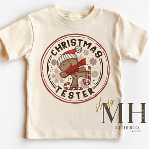 May include: Cream-colored t-shirt featuring a circular graphic with a gingerbread man wearing a Santa hat and scarf, holding a candy cane. The text "CHRISTMAS TESTER" is in a circular design. The shirt has short sleeves.