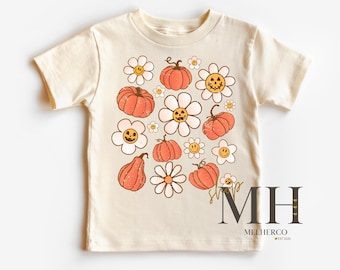 May include: Cream-colored t-shirt featuring a retro design of orange pumpkins and white daisies with smiling faces. The shirt has short sleeves and a crew neck. The design is a vintage-inspired print, perfect for fall or Halloween.