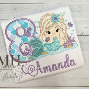 May include: A white t-shirt with a purple and teal number 8, a mermaid with a teal tail and blonde hair, and the name "Amanda" embroidered in purple.