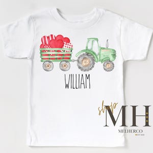 May include: White t-shirt with a watercolour illustration of a green tractor pulling a wagon filled with red and patterned hearts. The name "WILLIAM" is printed in black. The shop logo "MH" is in the bottom right corner.