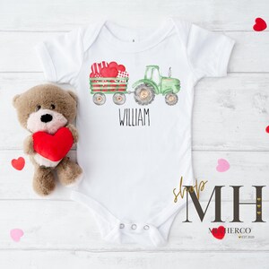 May include: White baby bodysuit featuring a green tractor and a wagon loaded with heart-shaped gifts. The name "WILLIAM" is printed below the tractor. A teddy bear holding a red heart is placed beside the bodysuit.
