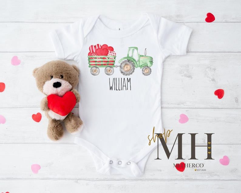 May include: White baby bodysuit featuring a green tractor and a wagon loaded with heart-shaped gifts. The name "WILLIAM" is printed below the tractor. A teddy bear holding a red heart is placed beside the bodysuit.