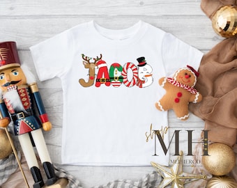May include: White short-sleeved t-shirt with the name "Jacob" in a festive Christmas design. The letters are decorated with a reindeer, candy cane, snowman, and elf. A gingerbread man and Christmas ornaments are also visible.