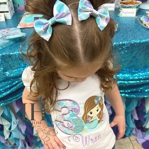 May include: A young girl wearing a white shirt with a mermaid design and the name "Wren" embroidered on it. She is wearing two blue and purple hair bows.