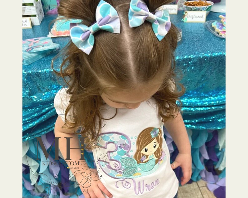 May include: A young girl wearing a white shirt with a mermaid design and the name "Wren" embroidered on it. She is wearing two blue and purple hair bows.