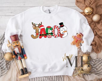 May include: White sweatshirt with the name "Jacob" in a festive Christmas design. The letters are decorated with a reindeer, Santa hat, elf, candy cane, and snowman. A gingerbread man and ornaments are also visible.