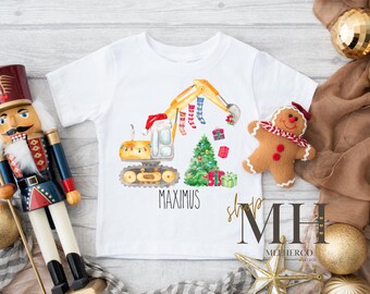 May include: White short-sleeve t-shirt featuring a Christmas-themed excavator design with a Santa hat, Christmas tree, and presents. The name "MAXIMUS" is printed below the design. A gingerbread man and nutcracker are in the background.