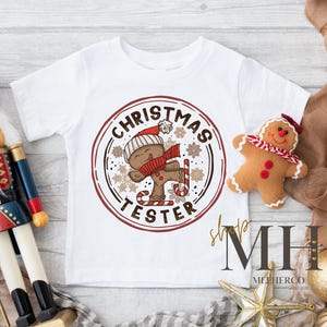 May include: White short-sleeved t-shirt featuring a circular design with a gingerbread man wearing a hat and scarf, holding a candy cane, and the text "CHRISTMAS TESTER". A gingerbread man plushie and a nutcracker are also visible.
