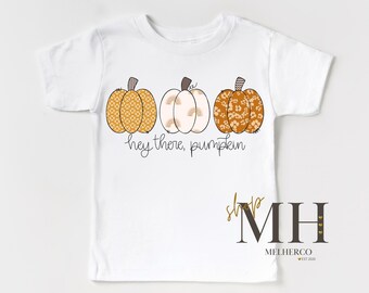 May include: White t-shirt featuring three pumpkins in shades of orange, cream, and yellow. The pumpkins have various patterns and textures. The text "hey there, pumpkin" is written in a cursive font. The shirt has a crew neck and short sleeves.