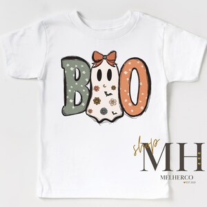K&ouml;nnte beinhalten: Wei&szlig;es T-Shirt mit einem Halloween-Design mit dem Wort "BOO". Das "B" ist gr&uuml;n mit wei&szlig;en Blumenakzenten, das "O" ist orange mit wei&szlig;en Punkten und das zweite "O" ist ein Geist mit einer Schleife und floralen Details.