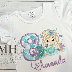 May include: White t-shirt with a purple and blue mermaid with a crown and a number 8. The mermaid is holding a seashell. The name Amanda is embroidered in purple below the mermaid.
