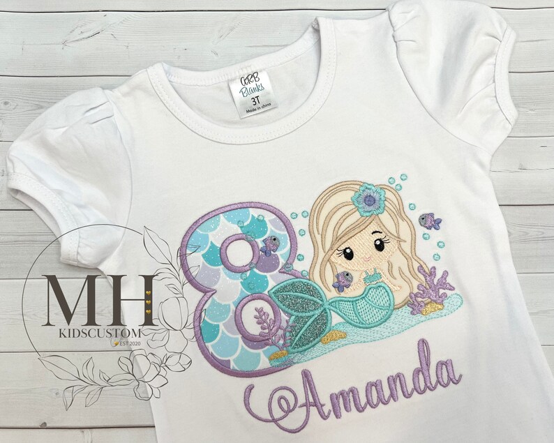 May include: White t-shirt with a purple and blue mermaid with a crown and a number 8. The mermaid is holding a seashell. The name Amanda is embroidered in purple below the mermaid.
