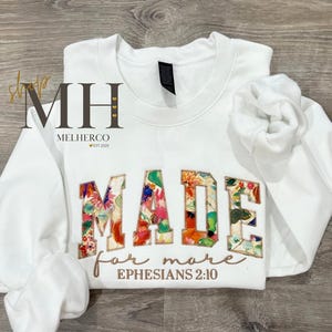May include: White sweatshirt with the word "MADE" in floral patchwork letters. Below, the words "for more" and "Ephesians 2:10" are embroidered in a brown script. The sleeves are folded, and the background is a wooden surface.