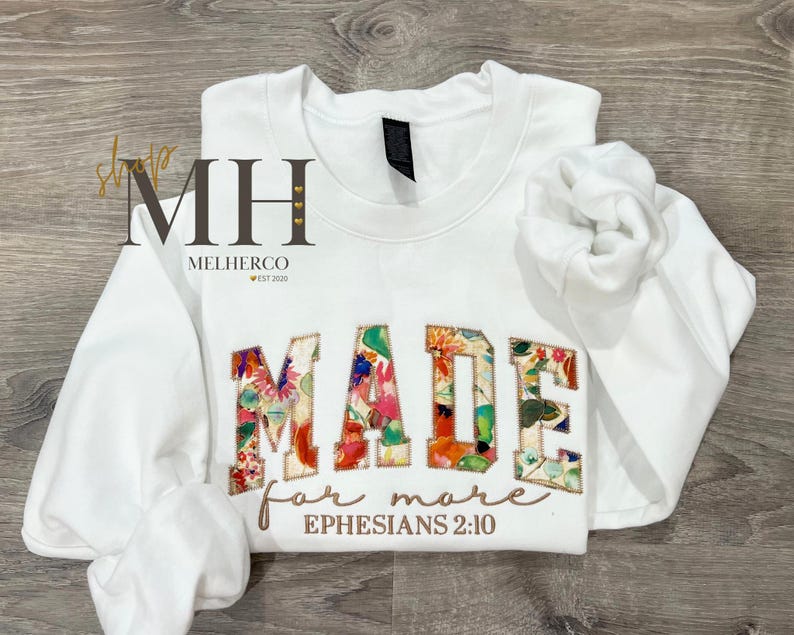 May include: White sweatshirt with the word "MADE" in floral patchwork letters. Below, the words "for more" and "Ephesians 2:10" are embroidered in a brown script. The sleeves are folded, and the background is a wooden surface.