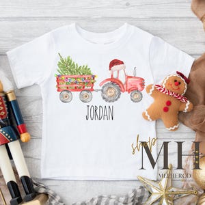 May include: White short-sleeved t-shirt featuring a watercolor illustration of a red tractor pulling a wagon with a Christmas tree and lights. The name "JORDAN" is printed below the graphic. A gingerbread man and nutcracker are also in the image.