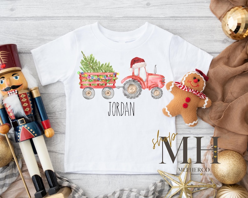 May include: White short-sleeved t-shirt featuring a watercolor illustration of a red tractor pulling a wagon with a Christmas tree and lights. The name "JORDAN" is printed below the graphic. A gingerbread man and nutcracker are also in the image.