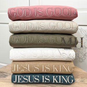 May include: Stack of six sweatshirts with embroidered text. The top two sweatshirts are pink and beige with the text "GOD IS GOOD". The next two sweatshirts are green and white with the text "GOD IS GOOD". The bottom two sweatshirts are brown and teal with the text "JESUS IS KING".