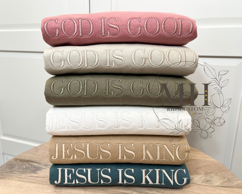May include: Stack of six sweatshirts with embroidered text. The top two sweatshirts are pink and beige with the text "GOD IS GOOD". The next two sweatshirts are green and white with the text "GOD IS GOOD". The bottom two sweatshirts are brown and teal with the text "JESUS IS KING".