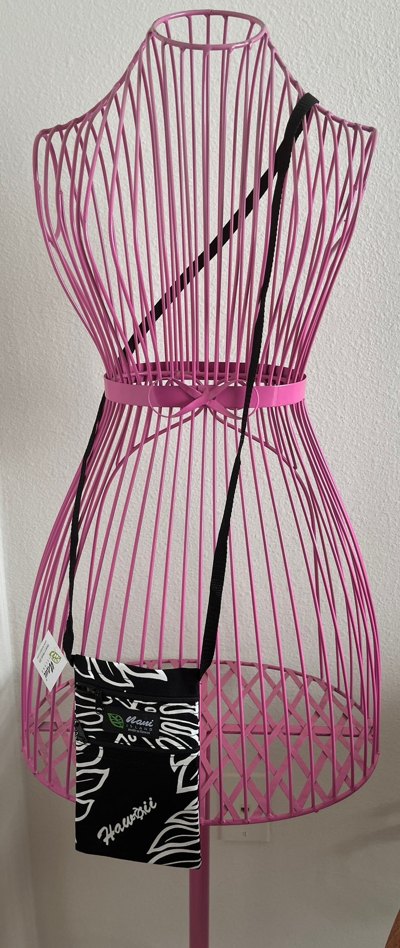 May include: A black and white crossbody bag with a floral design and the word "Hawaii" printed on it. The bag has a black strap and is displayed on a pink wireframe mannequin.