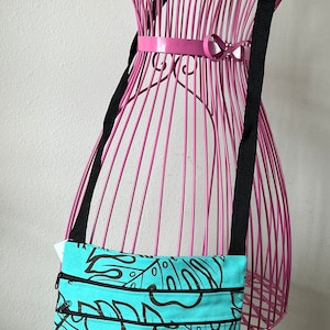 May include: A turquoise blue crossbody bag with a black strap and a floral print. The bag has two zippered compartments and a small label that reads "Maui Island Aloha Made in Hawaii".