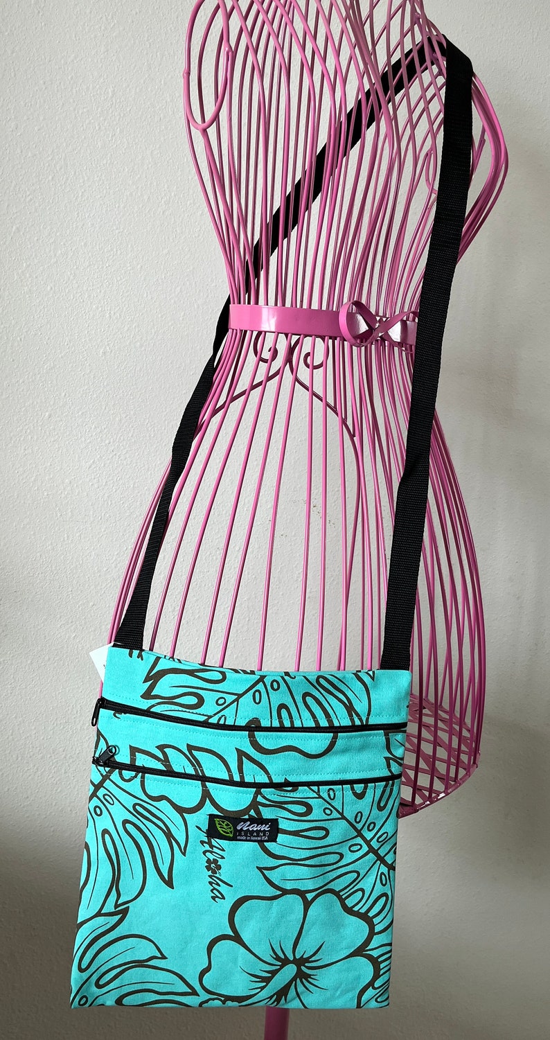 May include: A turquoise blue crossbody bag with a black strap and a floral print. The bag has two zippered compartments and a small label that reads "Maui Island Aloha Made in Hawaii".