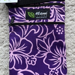 May include: A purple and light pink floral print crossbody bag with a black zipper and strap. The bag has a label that says "Nani Island made in Hawaii USA". The bag is made of a durable fabric.