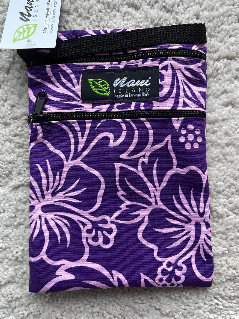May include: A purple and light pink floral print crossbody bag with a black zipper and strap. The bag has a label that says "Nani Island made in Hawaii USA". The bag is made of a durable fabric.