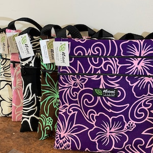 May include: A set of eight zippered pouches with floral patterns. The pouches are made of fabric and have a black zipper. The pouches are lined up in a row, with the largest pouch at the back and the smallest pouch at the front. The pouches are all different colors, including purple, pink, black, green, and white. The pouches have the text "Maui Island" printed on them.