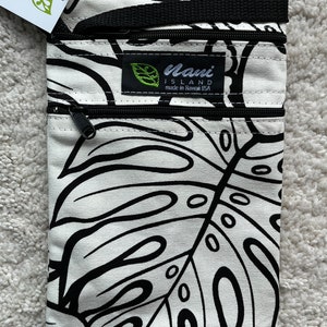 May include: A white and black crossbody bag with a tropical leaf pattern. The bag has two zippered compartments and a black strap. The label reads "Nani Island made in Hawaii USA".