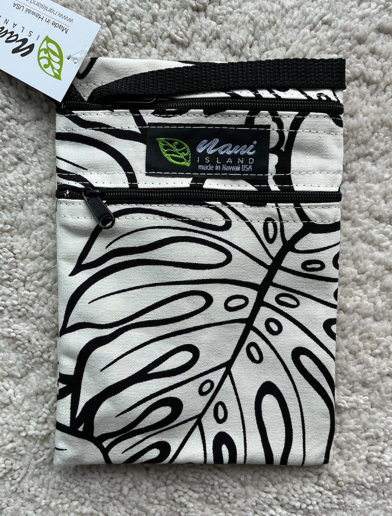 May include: A white and black crossbody bag with a tropical leaf pattern. The bag has two zippered compartments and a black strap. The label reads "Nani Island made in Hawaii USA".