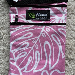 May include: Pink and white patterned crossbody bag with two zippered compartments. The bag features a black strap and zippers. The design includes large white leaf shapes on a pink background. The label reads "Maui Island made in Hawaii USA".