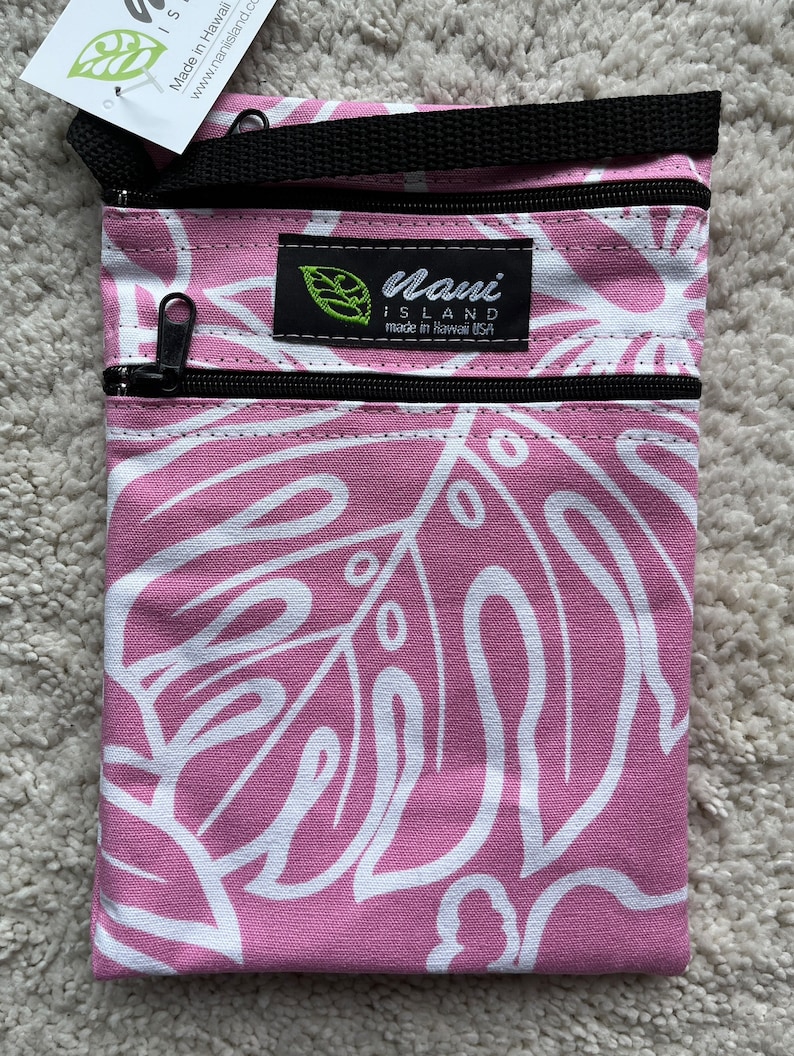 May include: Pink and white patterned crossbody bag with two zippered compartments. The bag features a black strap and zippers. The design includes large white leaf shapes on a pink background. The label reads "Maui Island made in Hawaii USA".