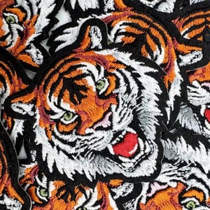 Könnte beinhalten: Eine Sammlung gestickter Tigerkopf-Aufnäher mit orangefarbenem, weißem und schwarzem Garn. Jeder Aufnäher zeigt einen Tiger mit grünen Augen, einer rosa Nase und einem geöffneten Maul, das weiße Zähne und eine leuchtend rote Zunge zeigt.