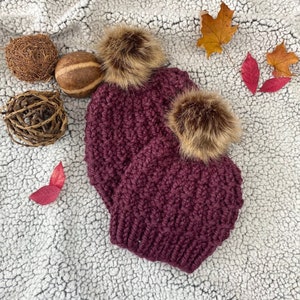 May include: Two burgundy knit beanies with faux fur pom poms. The beanies are on a white fuzzy surface with fall leaves.