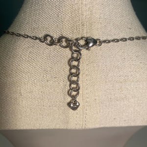 May include: Close-up of the back of a silver-toned necklace. The necklace features a chain with a lobster clasp and an adjustable chain with a small heart-shaped charm. The necklace is displayed on a beige fabric.