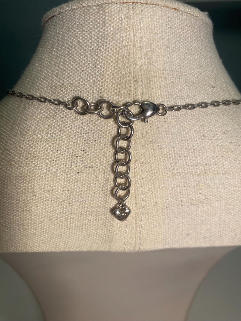 May include: Close-up of the back of a silver-toned necklace. The necklace features a chain with a lobster clasp and an adjustable chain with a small heart-shaped charm. The necklace is displayed on a beige fabric.