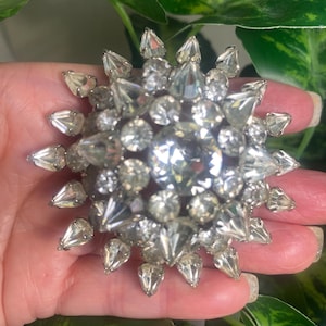 May include: A silver-toned, starburst-shaped brooch with clear, faceted crystals. The brooch features a large, round central stone surrounded by various sizes and shapes of crystals, creating a sparkling, eye-catching design. The brooch is a vintage accessory.