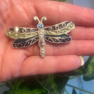 May include: A silver dragonfly brooch with green, black, and blue enamel. The dragonfly has clear and pink rhinestones on its body.