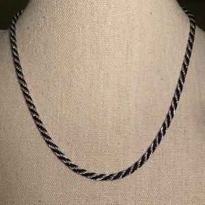 May include: A silver and black twisted rope necklace displayed on a mannequin. The necklace is made of interwoven strands of silver and black metal, creating a textured, braided appearance. The chain is a classic design.