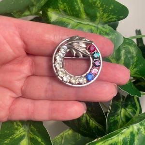 May include: A silver-toned brooch with a circular design. The brooch features a ring of clear and colorful gemstones. The gemstones include clear, red, green, blue, and pink stones. The brooch has a decorative leaf design.