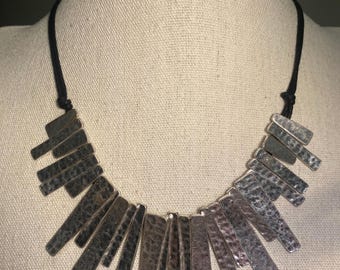 May include: A silver necklace with a hammered metal pendant. The pendant is made up of multiple rectangular pieces that are arranged in a fan shape. The necklace is on a black cord.
