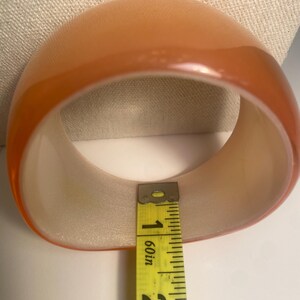 May include: A translucent orange bangle bracelet. The bracelet is measured with a yellow tape measure, showing a width of approximately 4.4 cm. The bracelet has a rounded, smooth exterior and a lighter interior.