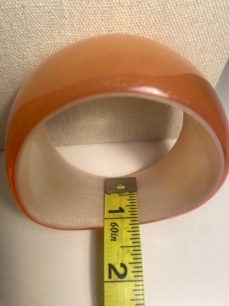 May include: A translucent orange bangle bracelet. The bracelet is measured with a yellow tape measure, showing a width of approximately 4.4 cm. The bracelet has a rounded, smooth exterior and a lighter interior.