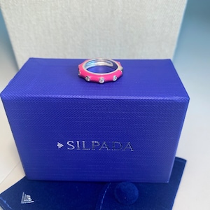 May include: A pink and silver ring with small clear stones set in the band. The ring is displayed in a blue velvet box with the brand name "SILPADA" printed on the lid.