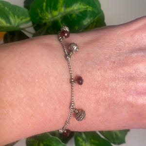 K&ouml;nnte beinhalten: Silberfarbenes Charm-Armband mit h&auml;ngenden dekorativen Elementen. Das Armband hat kleine, runde, rot gef&auml;rbte Perlen und strukturierte Silberanh&auml;nger. Das Armband wird vor einem Hintergrund aus gr&uuml;nen Bl&auml;ttern gezeigt.