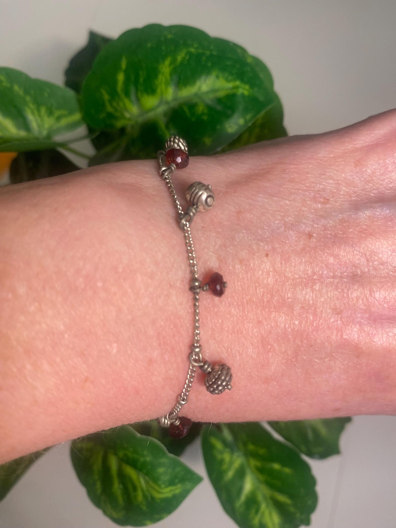 K&ouml;nnte beinhalten: Silberfarbenes Charm-Armband mit h&auml;ngenden dekorativen Elementen. Das Armband hat kleine, runde, rot gef&auml;rbte Perlen und strukturierte Silberanh&auml;nger. Das Armband wird vor einem Hintergrund aus gr&uuml;nen Bl&auml;ttern gezeigt.
