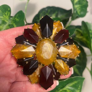 May include: A large brooch featuring a central oval amber-colored stone surrounded by a cluster of faceted, geometric-shaped stones in shades of deep brown, dark red, and marbled yellow, all set within a gold-toned metal frame.