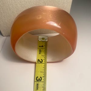 May include: A translucent orange bangle bracelet. The bracelet is measured with a yellow tape measure, showing an approximate width of 7.6 cm. The interior of the bracelet is a light beige colour.